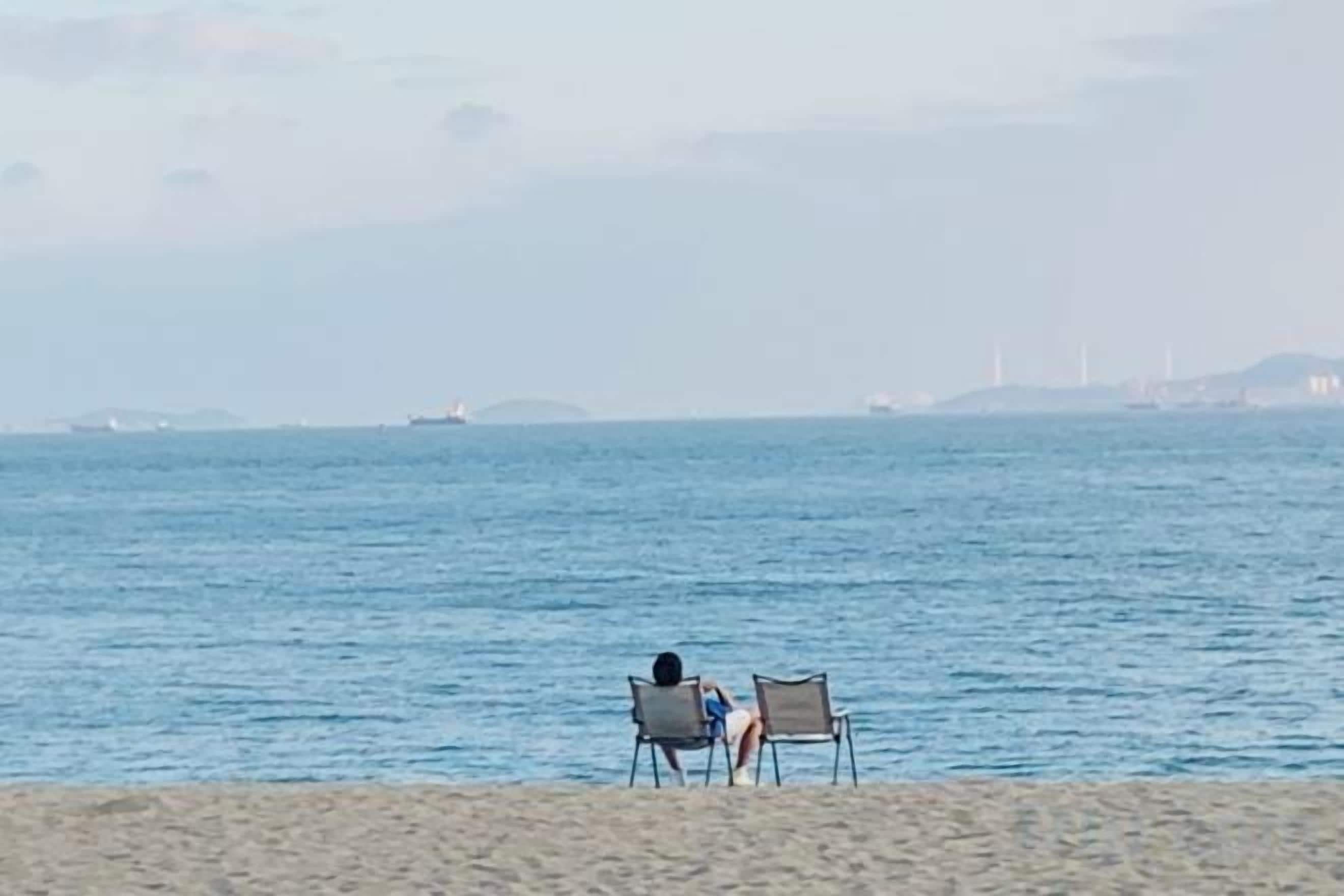 Una playa con dos sillas mirando al mar, un barco distante y un horizonte brumoso.