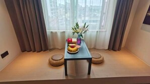 Quarto com uma plataforma de madeira baixa, uma mesa pequena com um vaso e uma travessa de frutas, e vista para edifícios através de cortinas leves.