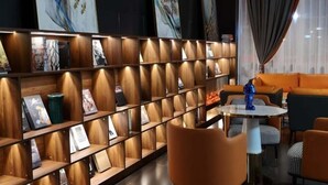 Um interior moderno com uma estante de madeira exibindo diversos objetos, uma mesa redonda com cadeiras e um sofá laranja.