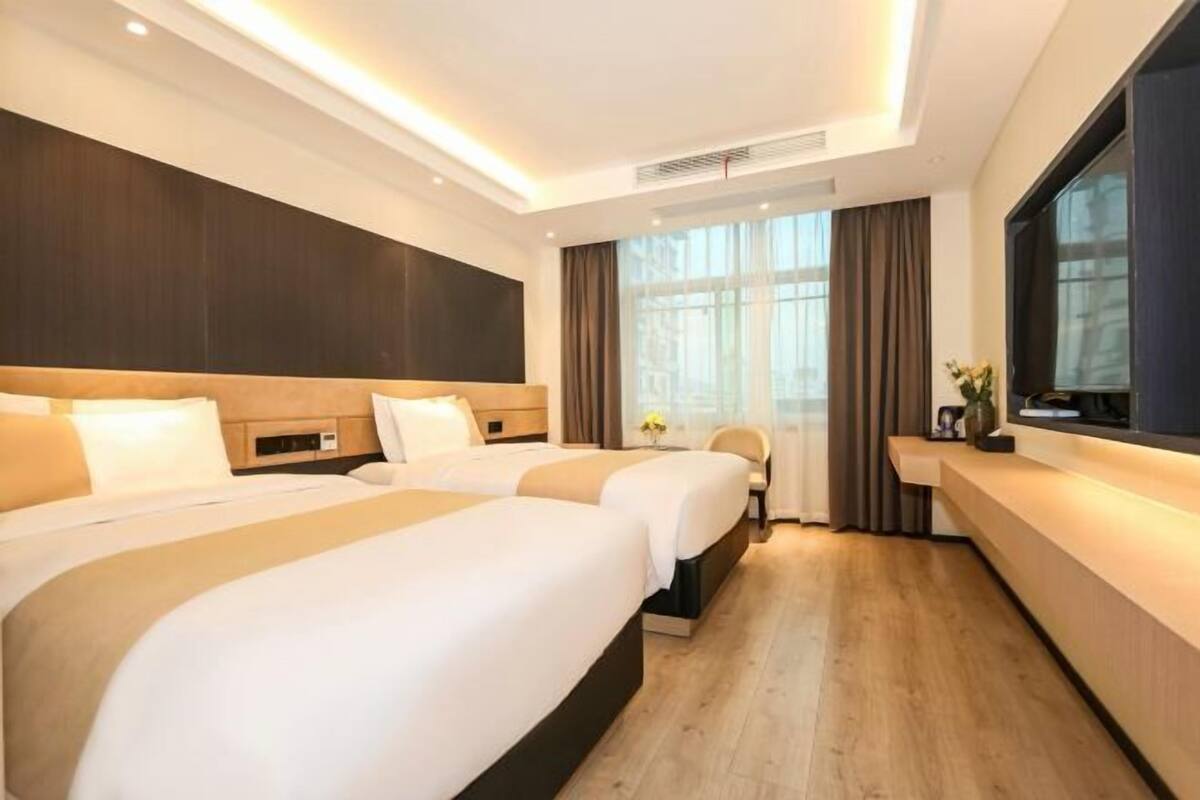 A hotel room with two beds, a desk, a TV, and a large window with curtains.