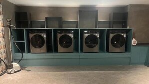 Un cuarto de lavandería con cuatro lavadoras en gabinetes verde azulado, una aspiradora y un pequeño bote de basura en el suelo.