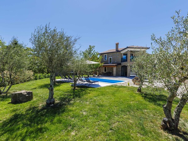Une maison en pierre à deux étages, dotée d’une piscine, entourée d’arbres et d’une pelouse verdoyante.
