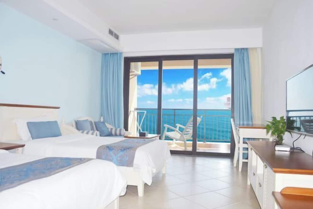 A hotel room with two beds, a balcony with a view of the sea, a desk, and a chair.
