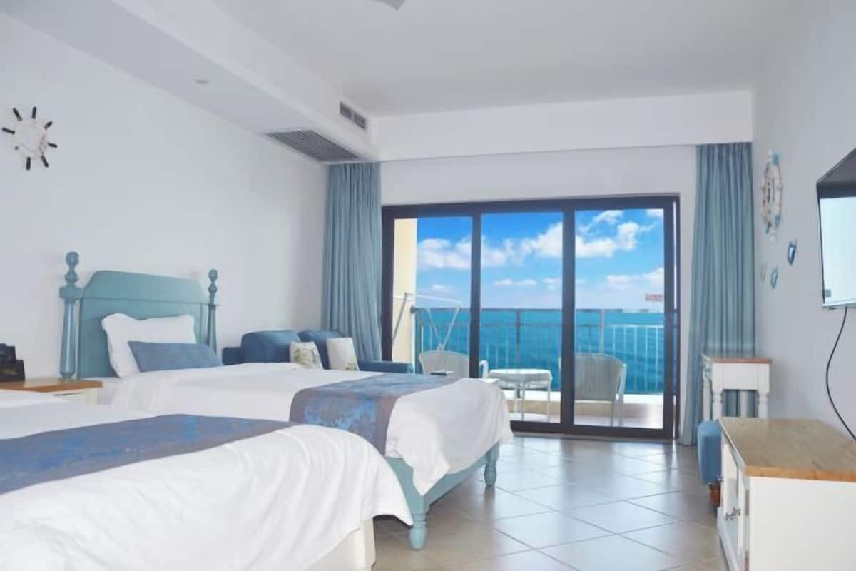 A hotel room with two beds, a balcony with a view of the sea, and a television on the wall.