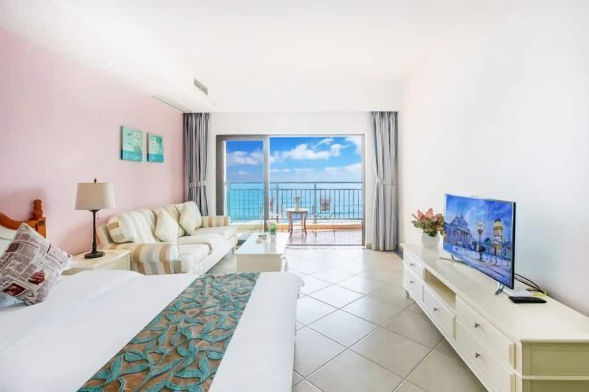 A hotel room with a bed, a sofa, a TV, a balcony with a view, and a dining area.