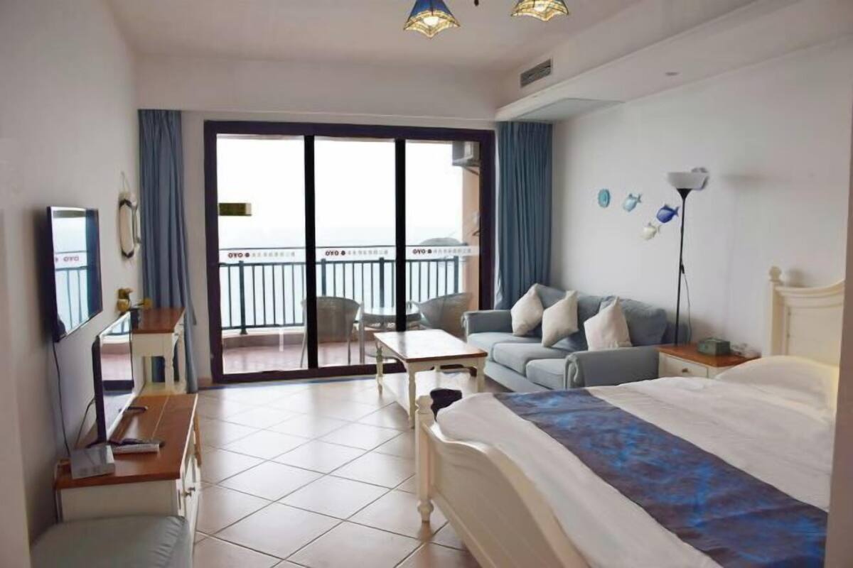 A hotel room with a bed, a desk, a chair, a sofa, a balcony with a railing, and a view of the outside.
