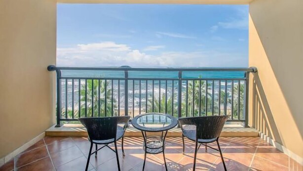 Un balcón con dos sillas de mimbre y una mesa de vidrio, con vista a una playa con palmeras y cielo despejado.