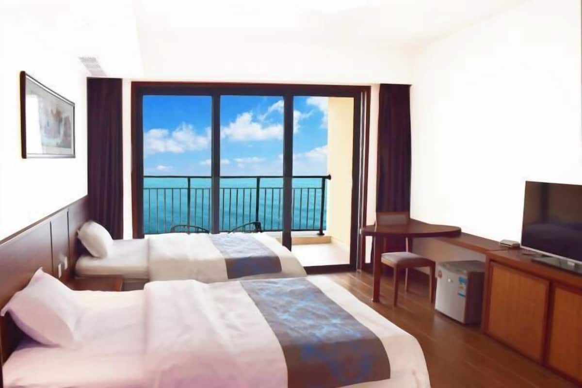 A hotel room with two beds, a desk, a TV, and a balcony with a view of the sea.