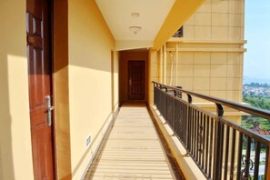 Un corridoio con una porta in legno a sinistra, un balcone con ringhiera nera a destra e vista su una cittadina in lontananza.