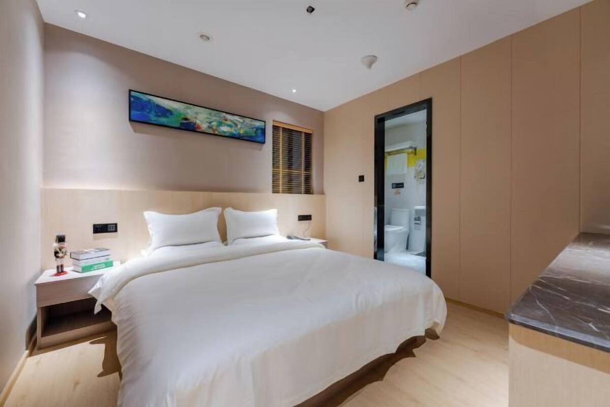 Une chambre moderne avec un grand lit, une table de chevet, un tableau au mur et une salle de bain visible à travers une porte en verre.