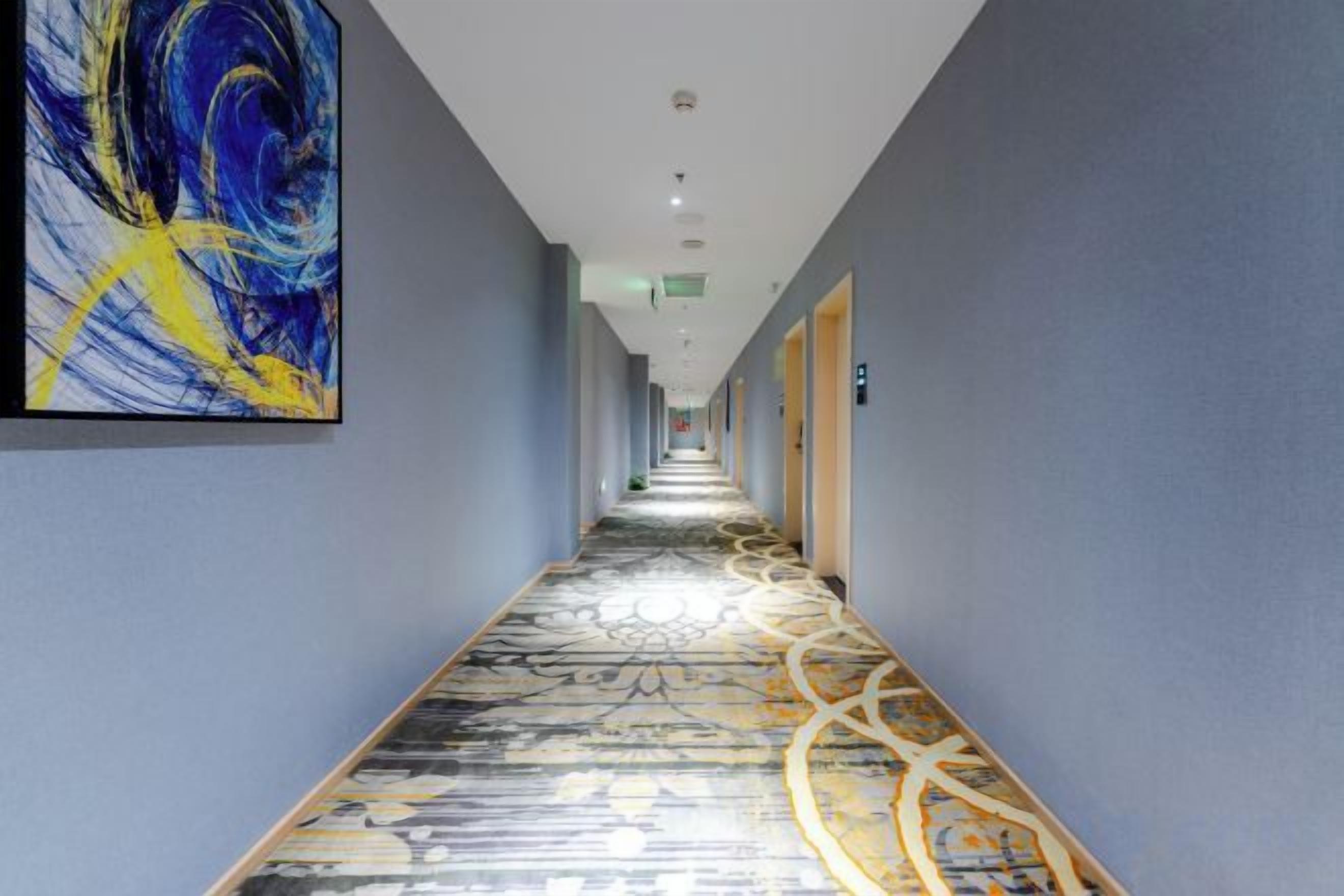 A hotel corridor with a patterned carpet, a large abstract painting on the left wall, and doors on the right.