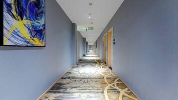 A hotel corridor with a patterned carpet, a large abstract painting on the left wall, and doors on the right.