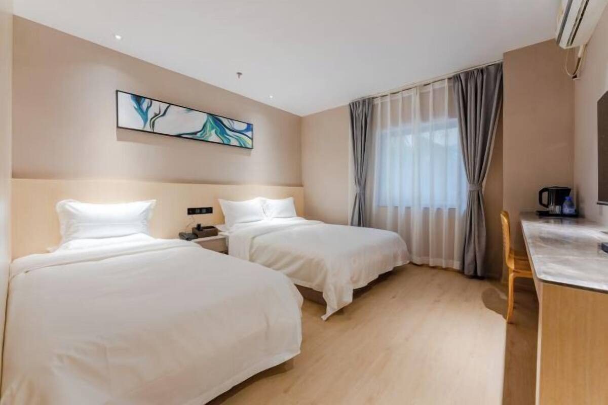 Une chambre d’hôtel avec deux lits, un bureau, une chaise, un tableau au mur et une fenêtre avec des rideaux.
