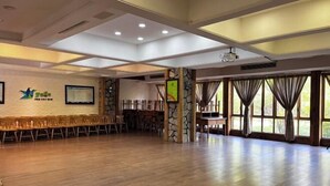 Ein geräumiger Saal mit Holzboden, einer Bühne mit Stühlen, einer Steinsäule und großen Fenstern mit Vorhängen.