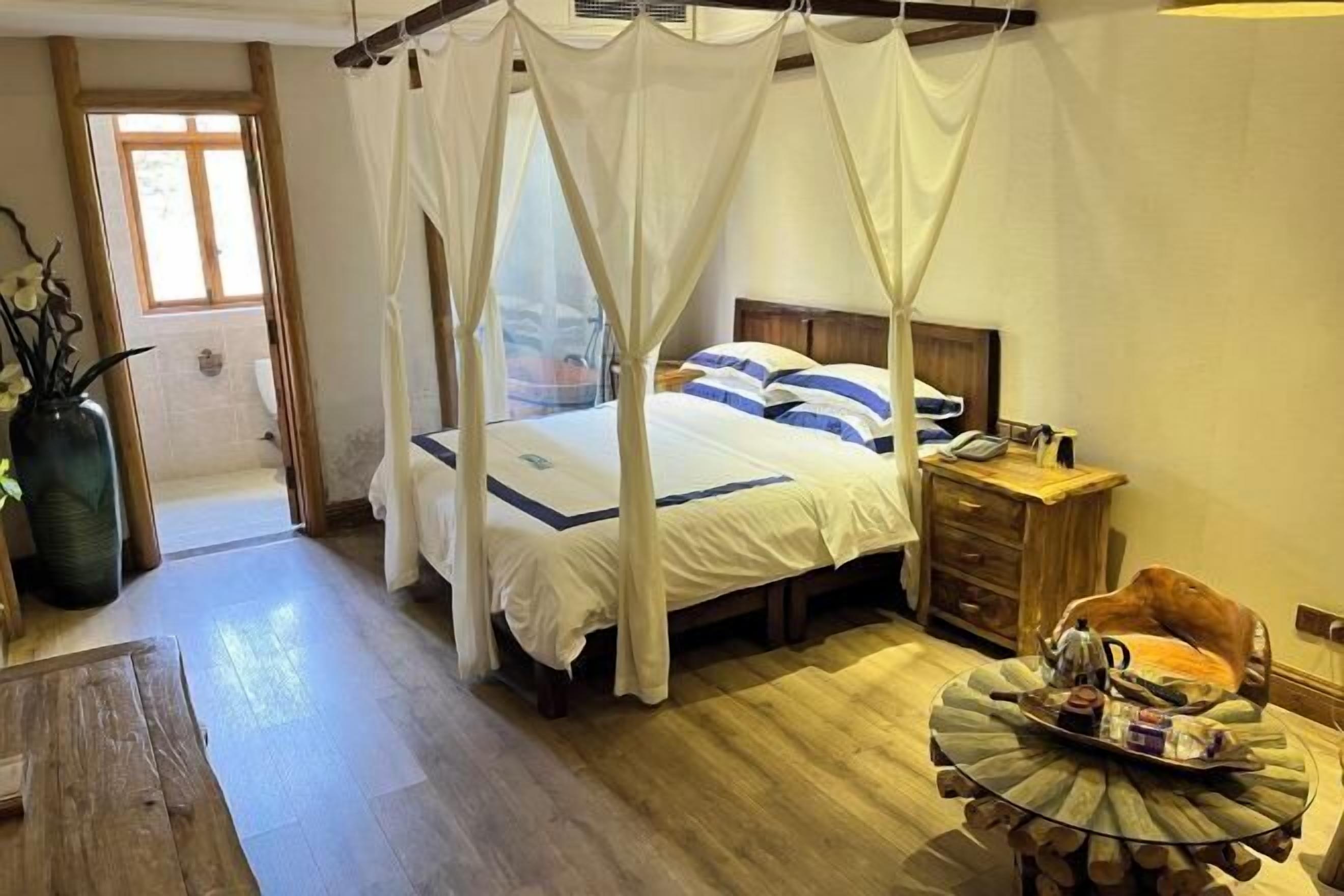 A bedroom with a canopy bed, a wooden nightstand, a round table with a tray, and a wooden floor.