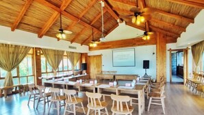Ein geräumiger Raum mit Holztischen und -stühlen, einer Projektionsleinwand und einem Deckenventilator.