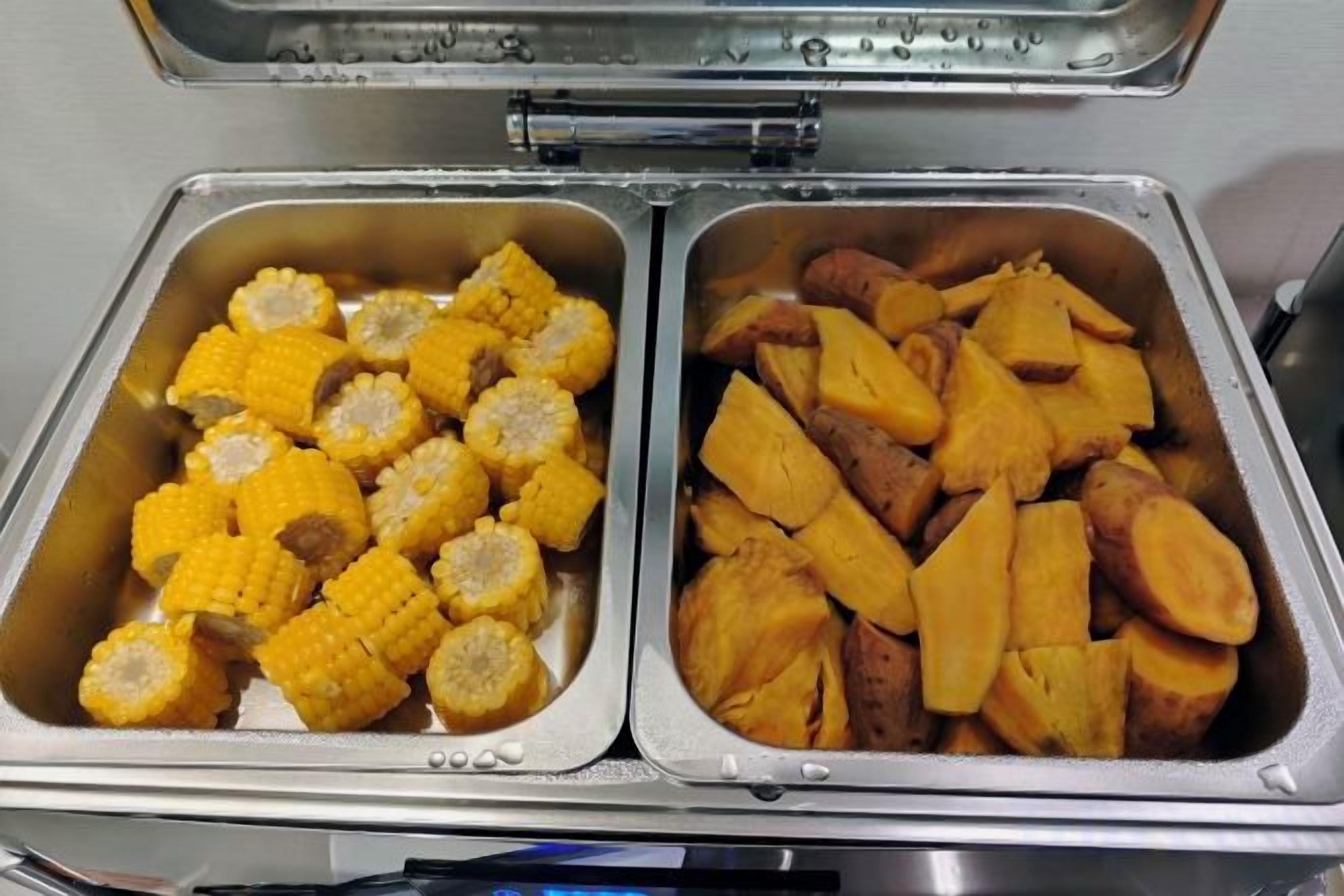 Un bufé con dos bandejas metálicas, una con mazorcas de maíz y la otra con batatas.