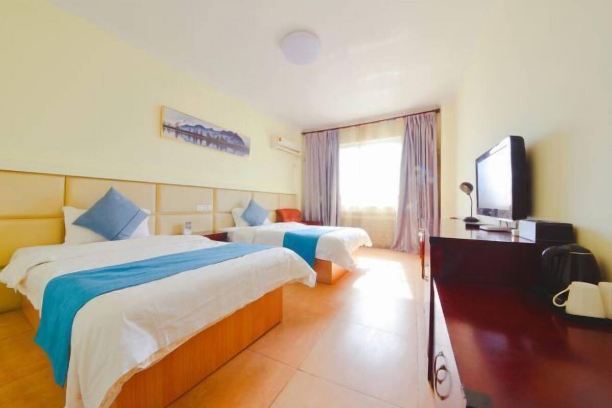 Ein Hotelzimmer mit zwei Betten, einem Flachbildfernseher, einem Schreibtisch mit Computer und einem Fenster mit Vorhängen.