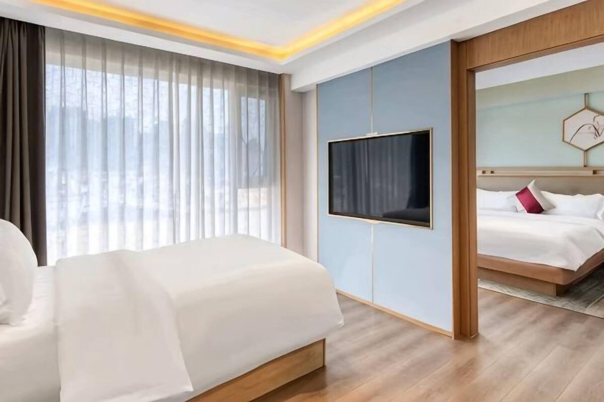 Ein Hotelzimmer mit einem großen Bett, einem an der Wand befestigten Fernseher und Blick auf ein weiteres Zimmer.