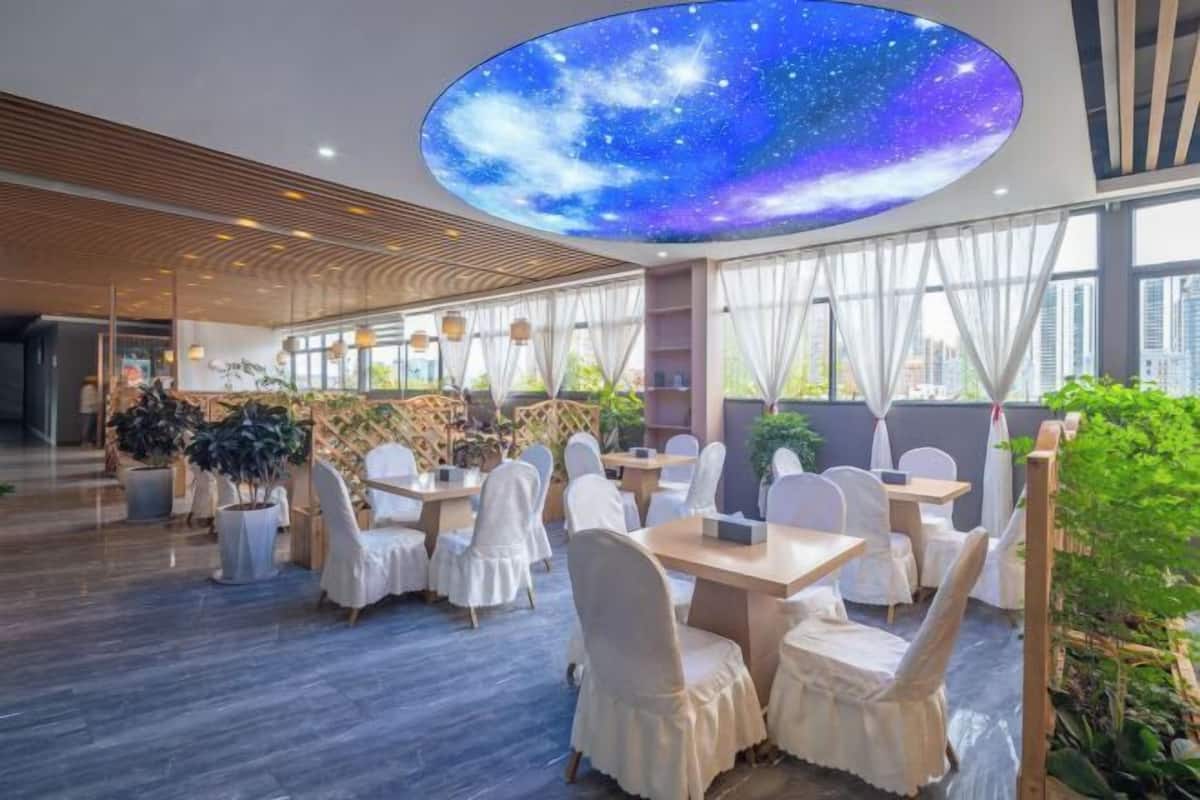 Uma sala de jantar moderna com mesas redondas e cadeiras brancas, um grande mural no teto com céu estrelado e amplas janelas que permitem entrada de luz natural.
