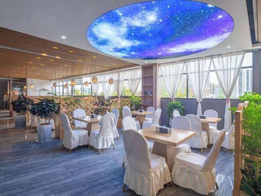 A modern dining area with round tables and white chairs, a large ceiling mural of a starry sky, and large windows allowing natural light.