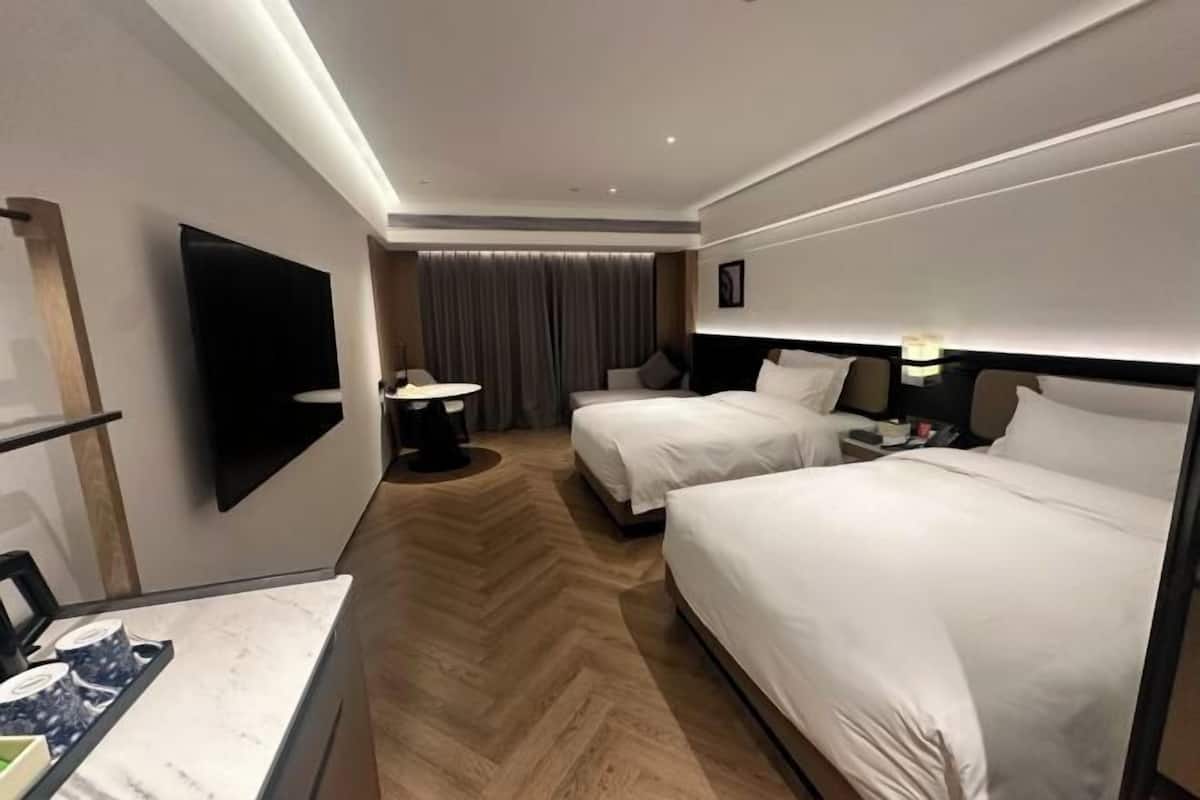 A hotel room with two beds, a flat-screen TV, a desk with a lamp, and a large window.