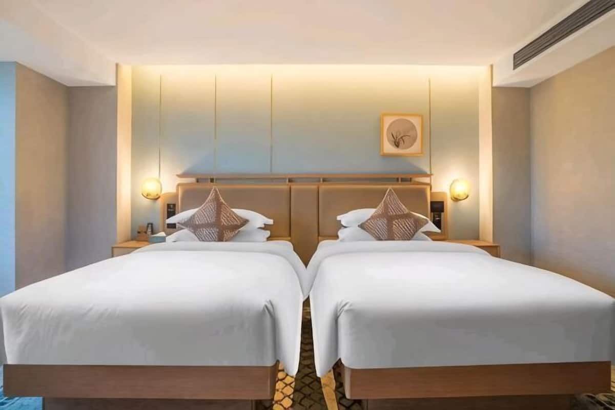 A hotel room with two beds, a headboard with integrated lighting, a framed picture on the wall, and a small bedside table with a lamp.