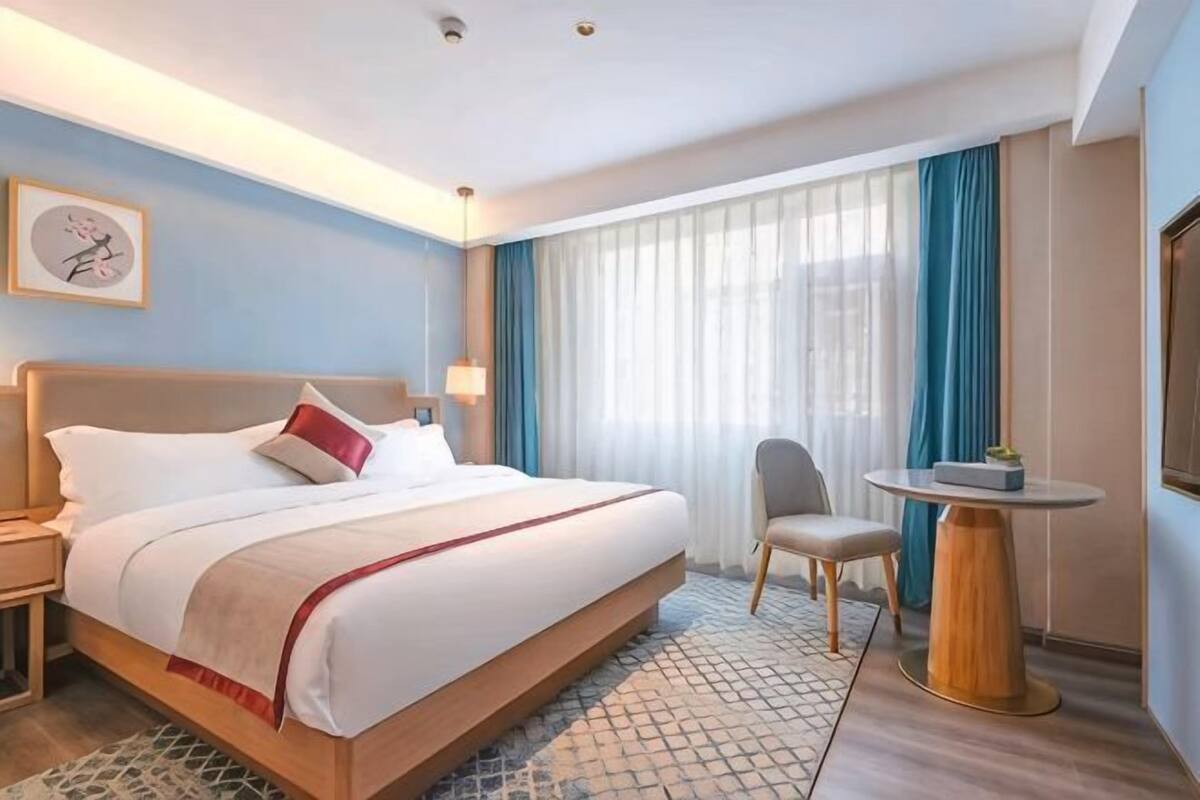 Ein Hotelzimmer mit einem großen Bett, einem Nachttisch, einem Sessel, einem kleinen runden Tisch und einem Fenster mit Vorhängen.
