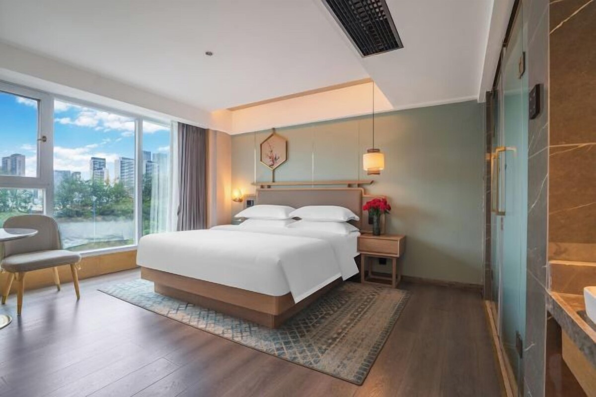 A modern hotel room with a large bed, a desk, a chair, and a view of the city.