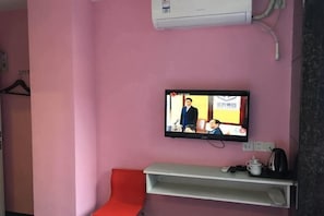 Una habitación rosada con una televisión instalada en la pared, una silla roja y un estante blanco con una tetera.