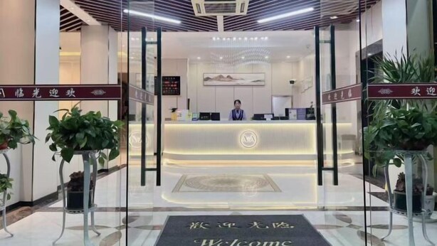 Zona de recepción de hotel con diseño moderno, que incluye un mostrador de recepción, un recepcionista y plantas en macetas.