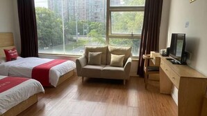 Habitación de hotel con dos camas, un sofá, un televisor y una ventana con vistas a edificios y árboles.