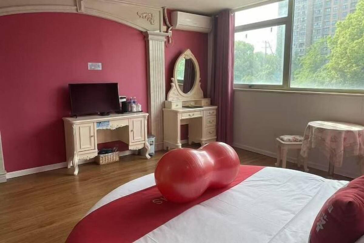 Une chambre d’hôtel équipée d’un lit, d’une télévision, d’une coiffeuse avec un miroir et d’une fenêtre donnant sur des immeubles et des arbres.