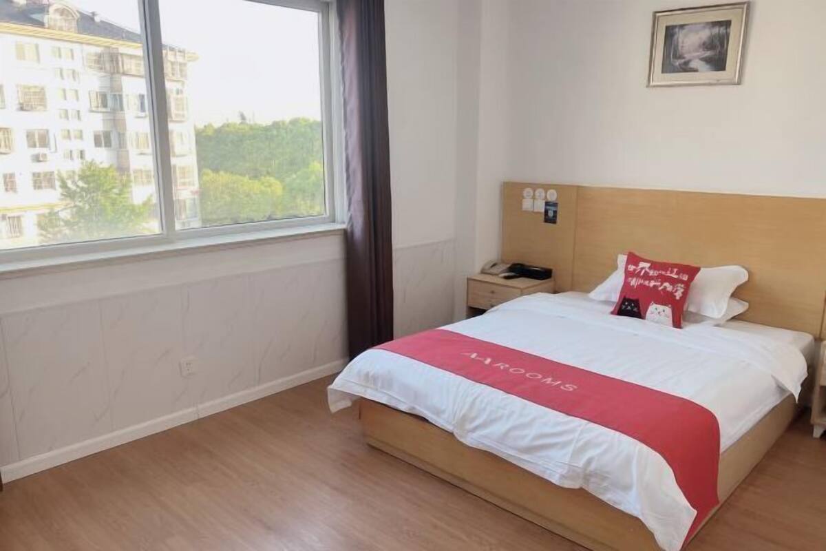 Une chambre d’hôtel avec un lit, une table de chevet, une fenêtre donnant sur des immeubles et un tableau accroché au mur.