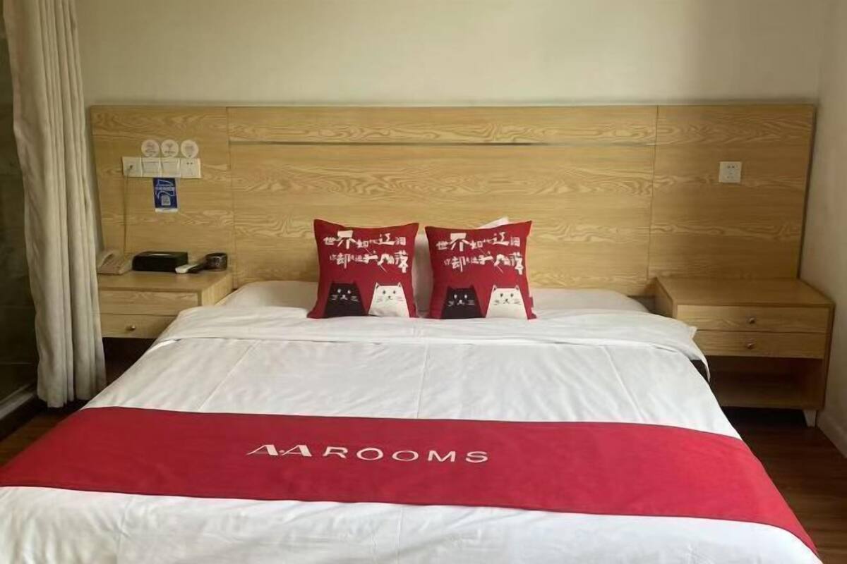 Une chambre d’hôtel avec une tête de lit en bois, un lit avec une literie blanche et rouge, et une couvre-lit rouge ornée du texte « AAROOMS ».