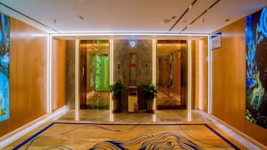 Un vestíbulo de hotel moderno con suelo de mármol, puertas de vidrio que dan a una zona de recepción y grandes cuadros abstractos en las paredes.