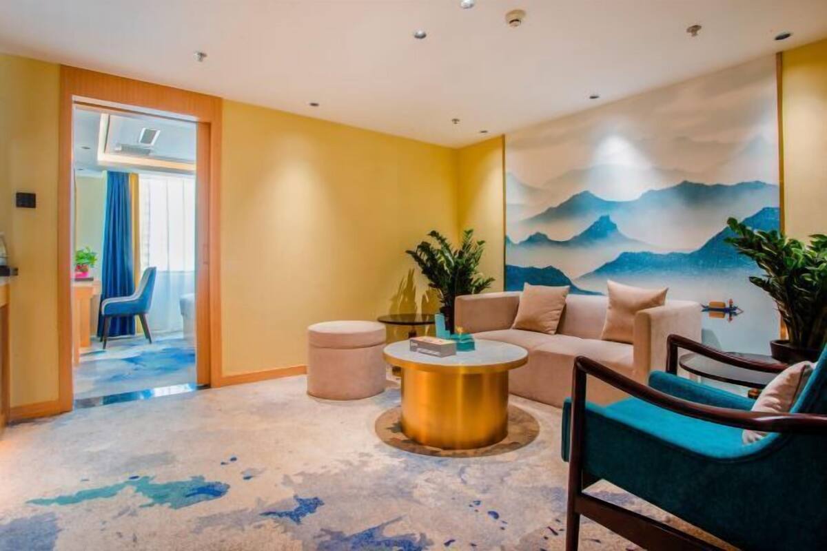 A modern living room with a beige sofa, a round coffee table, and a blue armchair. There is a large window with a view of mountains and a door leading to another room.