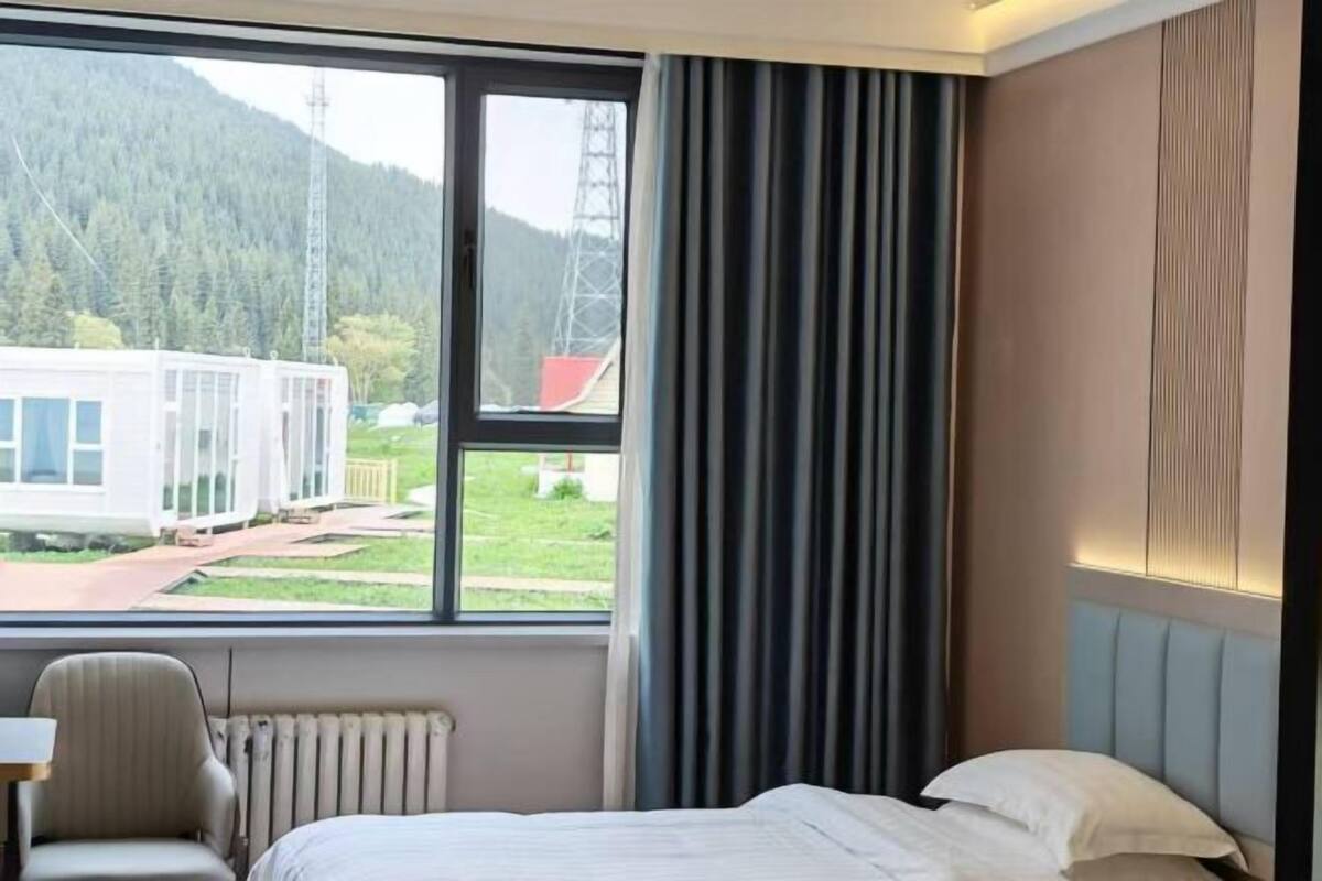 A hotel room with a bed, a chair, a window with curtains, and a view of a train and mountains.
