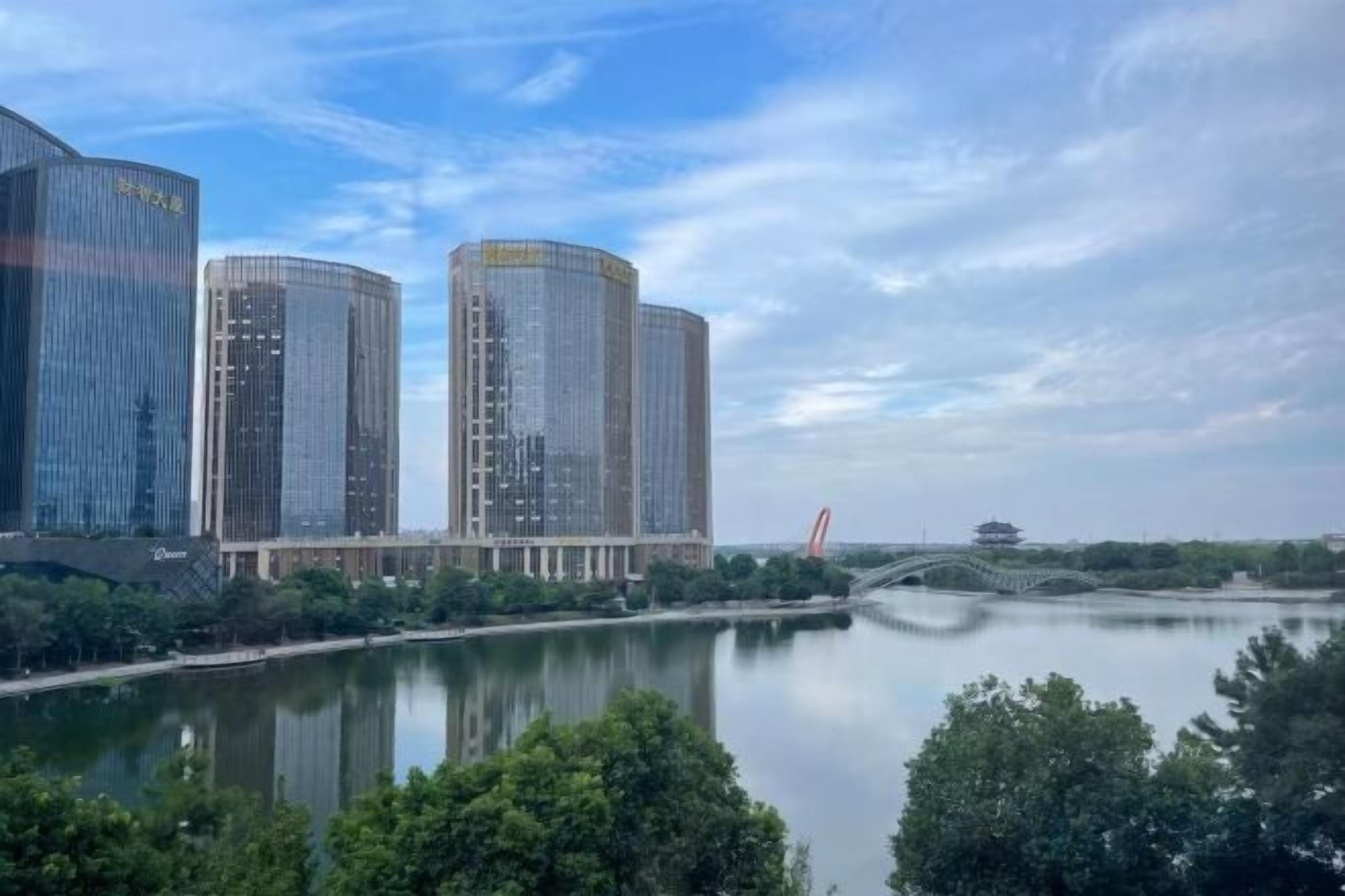 A river with modern buildings on the opposite bank, a clear sky, and a bridge in the distance.