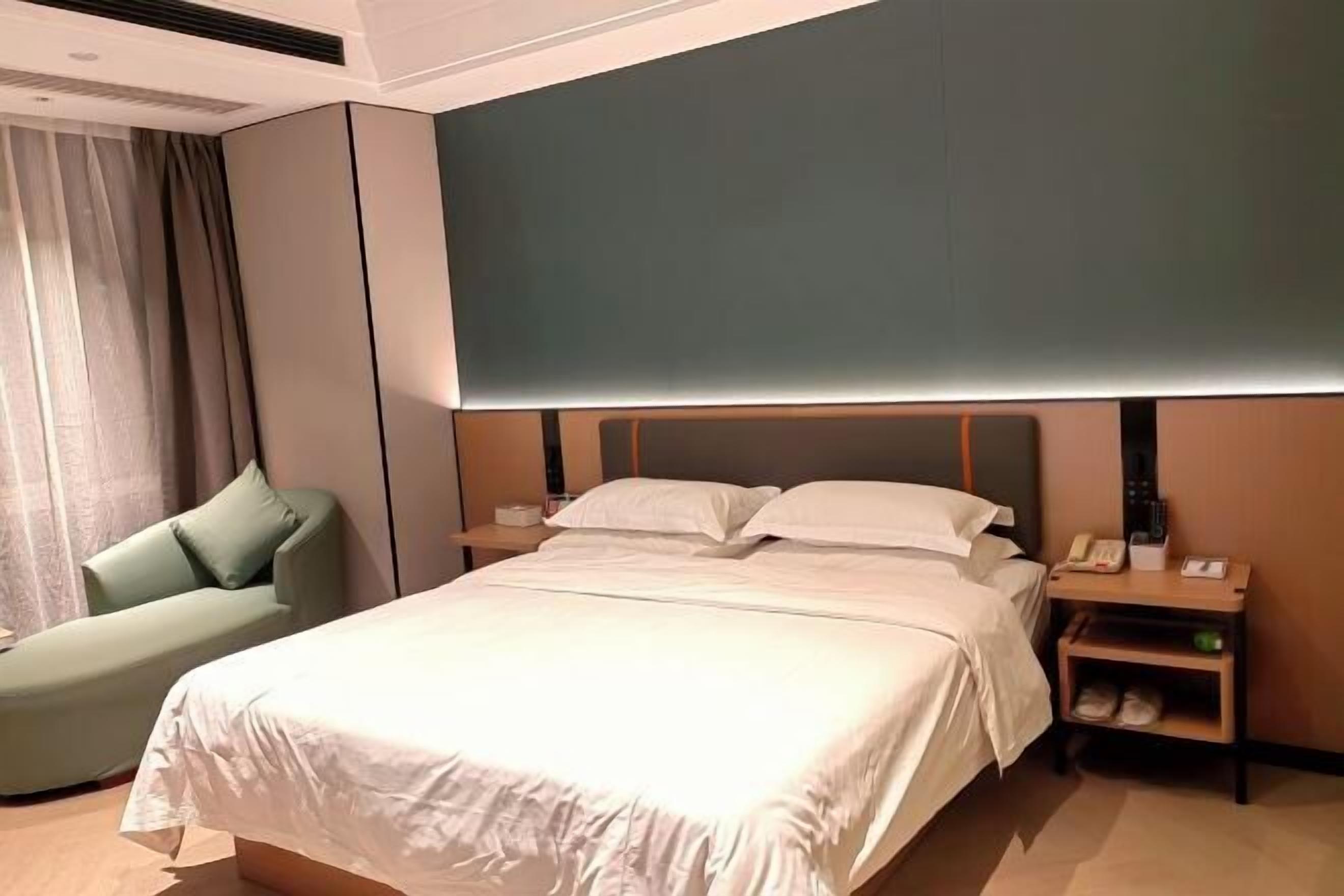 Ein modernes Hotelzimmer mit einem großen Bett, einem grünen Sessel, einem Nachttisch mit Lampe sowie einem Beistelltisch mit Telefon und einer kleinen Ablagefläche.