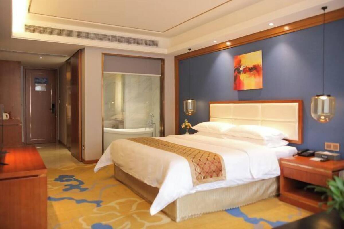 Une chambre d’hôtel avec un grand lit, une table de chevet, une commode, une lampe fixée au mur et un tableau.