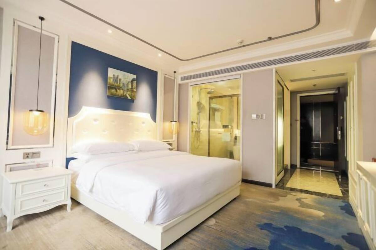 Une chambre d’hôtel moderne avec un grand lit, une table de chevet, une commode, une douche en verre et une vue sur le couloir.
