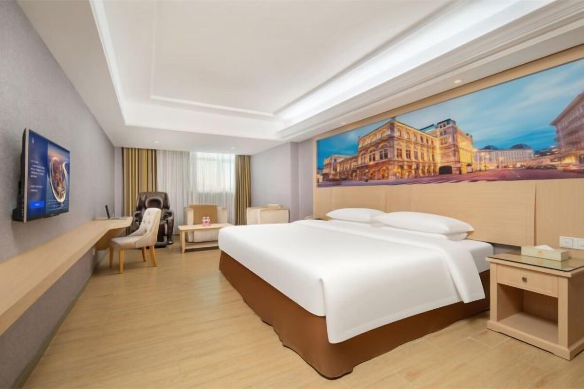 A hotel room with a large bed, a desk, a chair, and a painting of a cityscape.