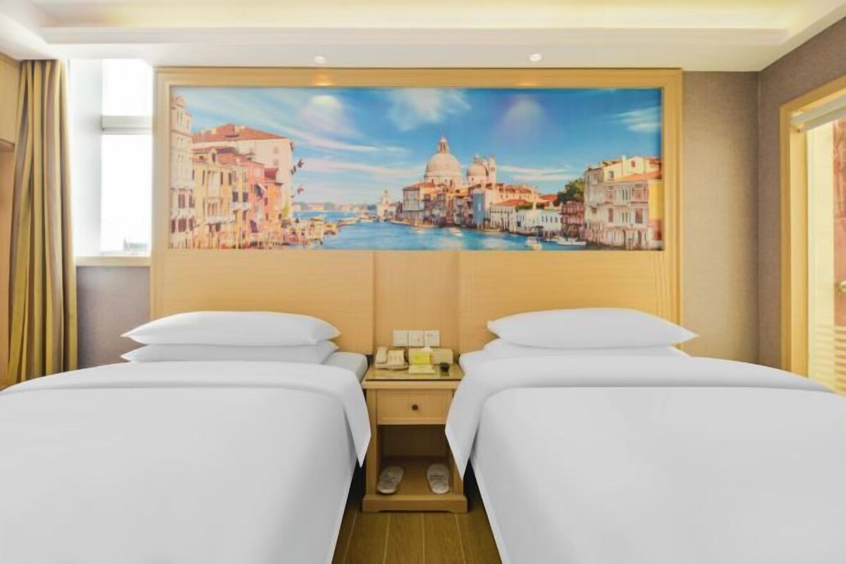 A hotel room with two beds, a painting of Venice, and a wooden nightstand with slippers.