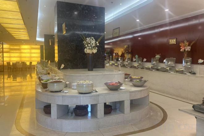 Vestíbulo de hotel con mostrador de recepción, una columna de mármol y una zona de bufé con diferentes recipientes de comida.