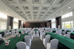 Un salón de conferencias con filas de sillas blancas, una larga mesa con manteles verdes y una pantalla grande al frente.
