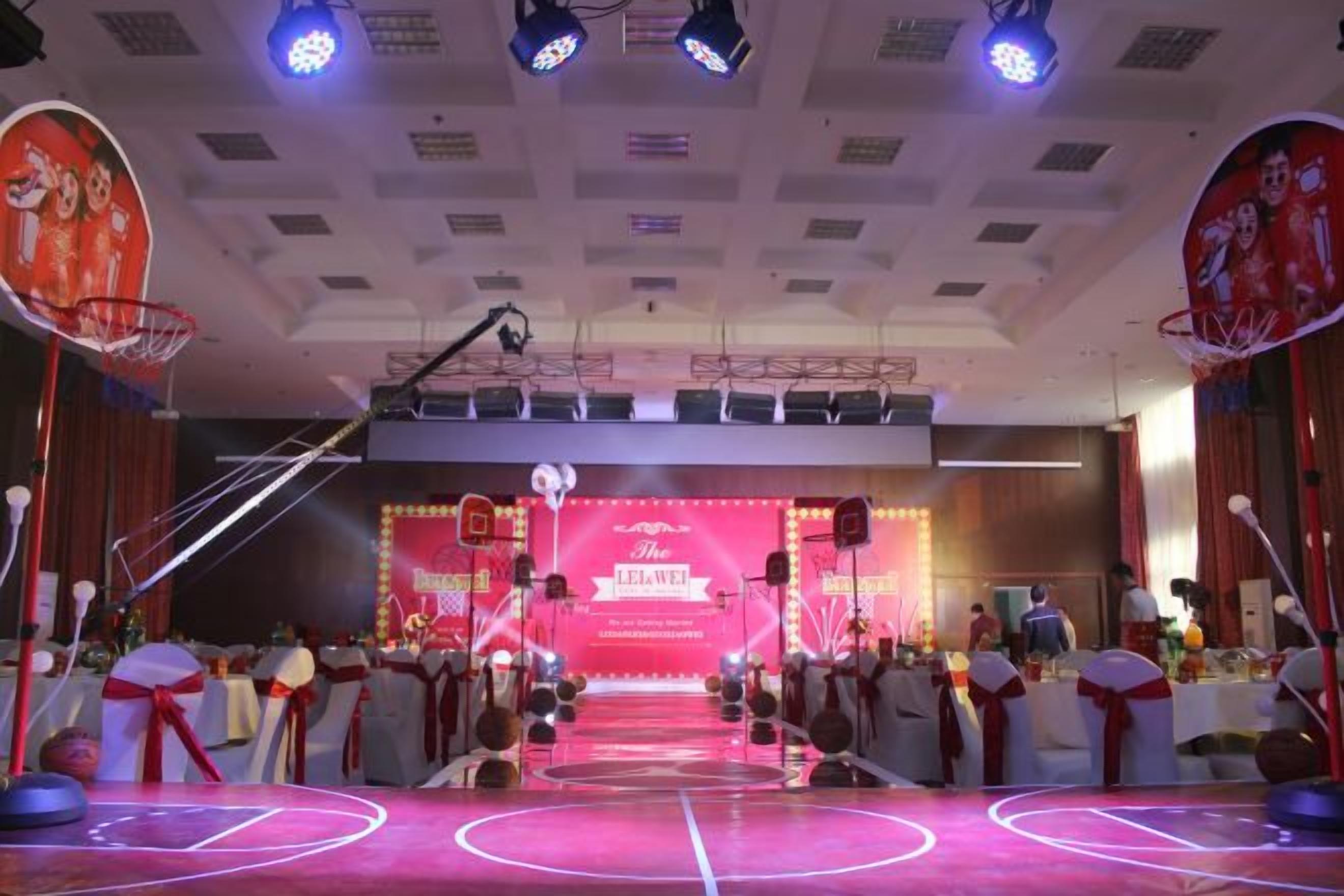An indoor basketball court with a stage in the background, audience seating, and stage lighting.