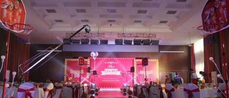 An indoor basketball court with a stage in the background, audience seating, and stage lighting.
