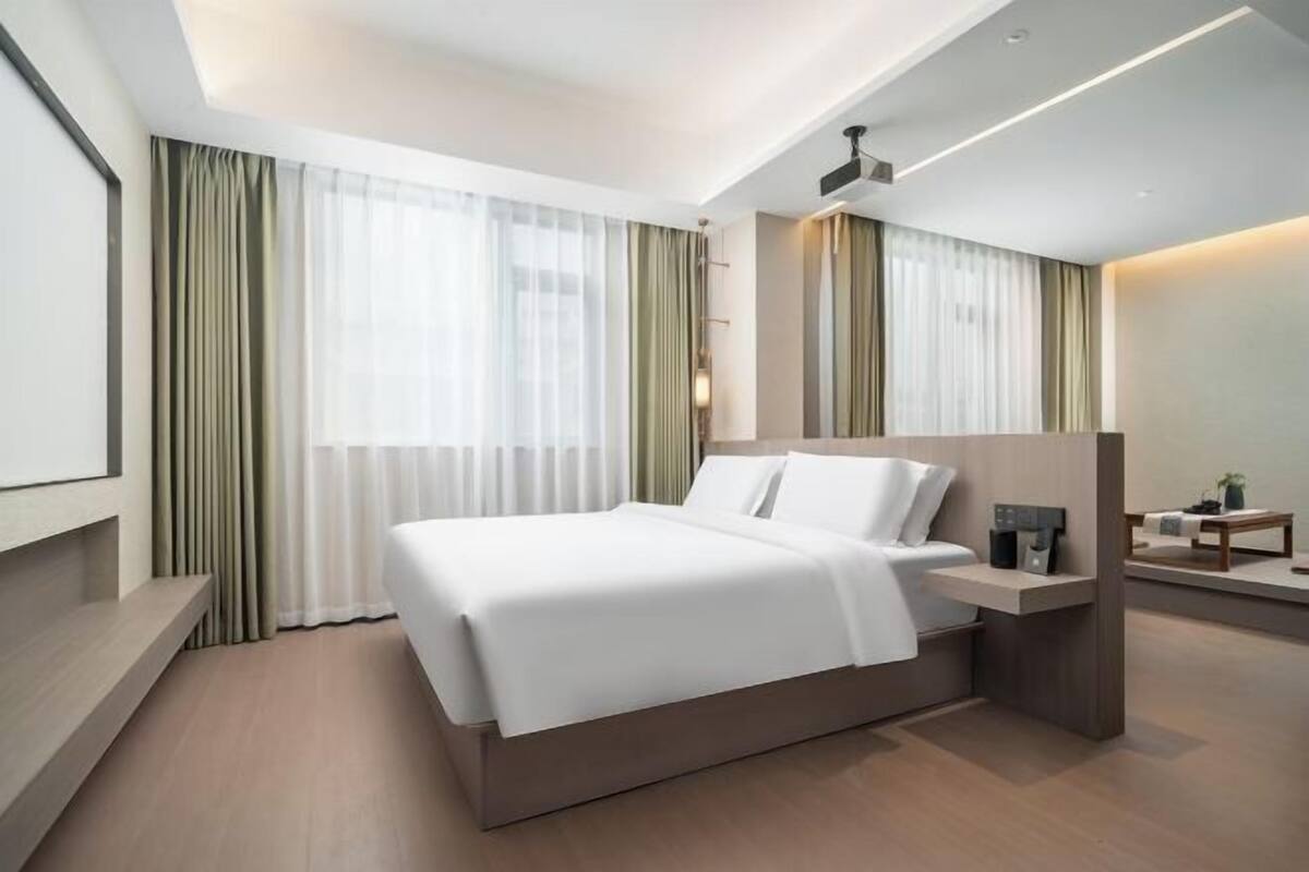 A modern hotel room with a large bed, a bedside table, a desk, and a chair.