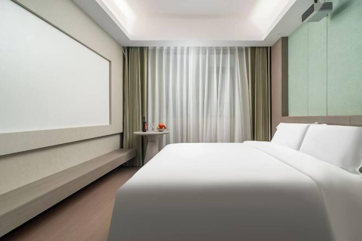 A modern hotel room with a bed, a small bedside table, a wall-mounted TV, and a window with curtains.
